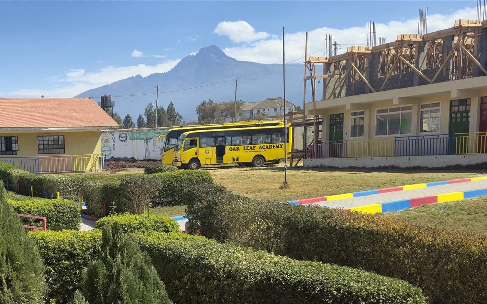 The yellow Oak Leaf Academy school bus parked on the campus lawn beside the classroom block, with Mount Kilimanjaro rising behind it.